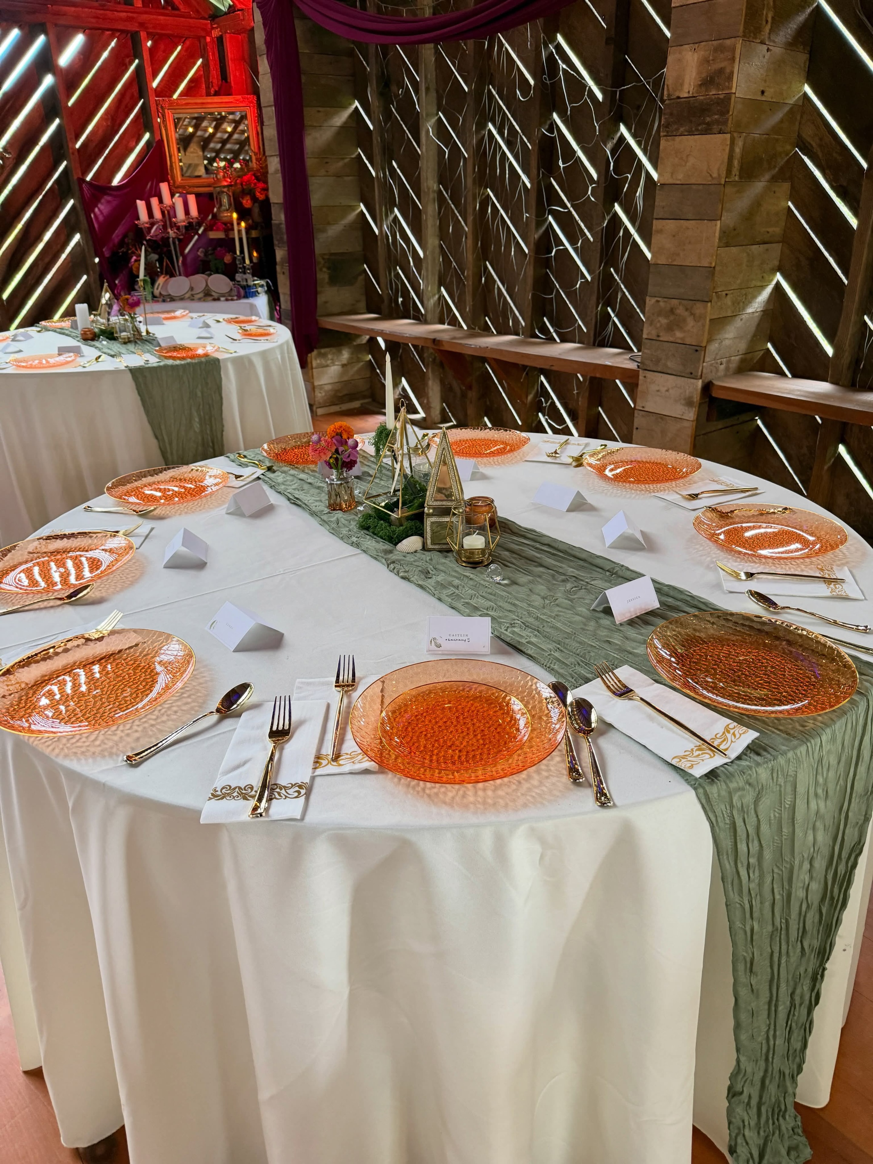 Round table with orange charger plates and sage green runner in a barn venue